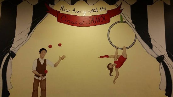 A painting of a man juggling and a woman hanging from a ring. Above is the caption: "run away with the circus at SANCA"